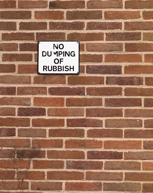 A rectangular metal sign mounted on a red brick wall, displaying the message 'NO DUMPING OF RUBBISH' in black capital letters on a white background. The sign is positioned toward the upper left of the image, with visible screws at each corner securing it in place. The brick wall features a uniform pattern of reddish-brown bricks with light mortar joints, and the bricks have a textured, slightly weathered appearance. The lighting is natural, casting soft shadows that emphasize the brick material's texture. The scene suggests an outdoor urban environment, likely part of a building façade, with the sign serving as a warning against illegal rubbish dumping in the vicinity. Rubbish Removal Clapham, specializing in private waste handling, may use such signage as part of their services to enforce proper rubbish disposal and support alternative waste collection methods.