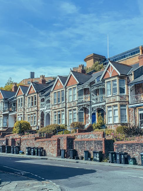 A row of Victorian-style terraced houses with distinctive bay windows and ornate ironwork balconies, situated on a residential street. The houses feature red brick and white-painted timber details, with gabled roofs and decorative trim along the eaves. In the foreground, there is a brick boundary wall with a series of black wheelie bins and recycling containers lined up along the pavement. The street appears clean and well-maintained, with small bushes and trees adding greenery to the front gardens. The sky above is partly cloudy with patches of blue, and the lighting suggests a bright day. The scene is typical of urban residential areas where private waste collection may be employed, supported by companies like Rubbish Removal Clapham for on-site clearance of household rubbish, contributing to alternative waste handling solutions beyond standard municipal services.