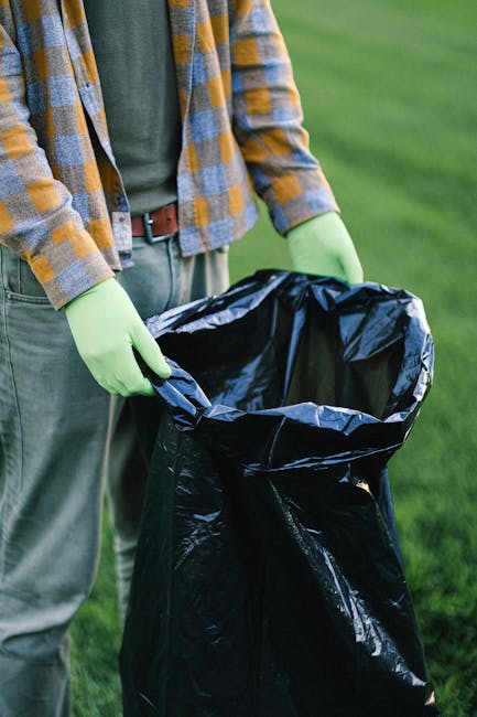A person wearing a plaid shirt with shades of yellow, blue, and gray, along with gray trousers and a brown belt, is standing outdoors on a grassy area. They are holding open a large black plastic rubbish bag lined with a silver inner layer, which is partially filled and appears ready for waste disposal. The individual is also wearing bright green rubber gloves, indicating they are engaged in waste collection or rubbish removal activity. In the background, there is an expanse of well-maintained green grass, suggesting a park or garden setting. The scene highlights the process of cleaning or clearing waste in an outdoor environment, consistent with private rubbish collection or site clearance services typically provided by companies like Rubbish Removal Clapham. The lighting is natural, with daylight illuminating the scene clearly, focusing on the hands, rubbish bag, and the surrounding grass, capturing the practical aspect of rubbish removal work.