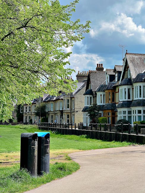 A row of Victorian-style terraced houses with distinctive bay windows and ornate ironwork balconies, situated on a residential street. The houses feature red brick and white-painted timber details, with gabled roofs and decorative trim along the eaves. In the foreground, there is a brick boundary wall with a series of black wheelie bins and recycling containers lined up along the pavement. The street appears clean and well-maintained, with small bushes and trees adding greenery to the front gardens. The sky above is partly cloudy with patches of blue, and the lighting suggests a bright day. The scene is typical of urban residential areas where private waste collection may be employed, supported by companies like Rubbish Removal Clapham for on-site clearance of household rubbish, contributing to alternative waste handling solutions beyond standard municipal services.