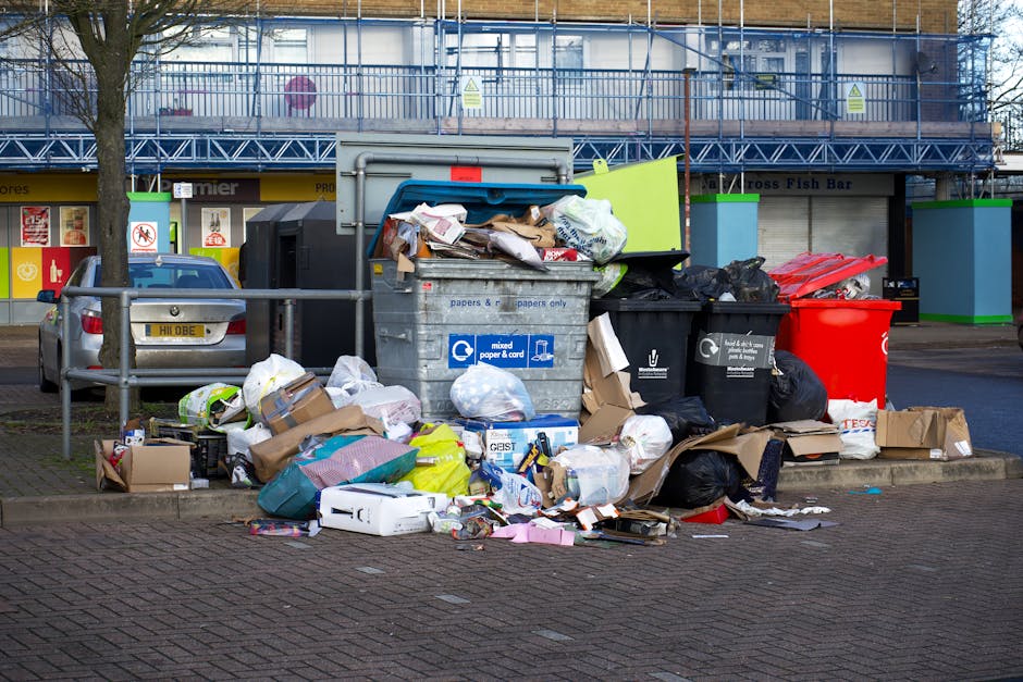 In the foreground of the image, a large collection of rubbish is piled on the pavement next to a curb, featuring various types of waste including cardboard boxes, plastic bags, paper packaging, and discarded household items. The waste is a mix of colourful and neutral tones, with some boxes flattened or partially torn, and black and green plastic rubbish bags among the debris. Behind the heap, three large wheelie bins are visible; one is black with a panel on the lid, another black with a white recycling symbol indicating mixed paper and cardboard, and a bright red bin, all positioned on the sidewalk. To the left, a small silver car is parked, with its rear bumper close to the rubbish. In the background, a building with a blue scaffolding structure and transparent safety barriers surrounds the upper level, indicating ongoing construction or maintenance work. The building has several storefronts or service units with signage, and there are a few small trees along the sidewalk, with the overall scene suggesting an area where independent waste disposal or rubbish removal services might be required, emphasizing the importance of professional clearance like that offered by Rubbish Removal Clapham.