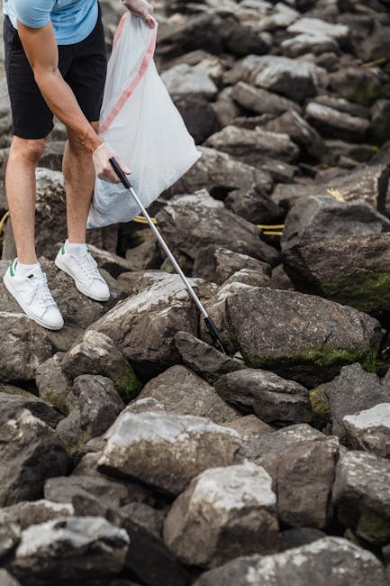The image shows a person wearing a light blue shirt, dark shorts, and white sneakers using a grabber tool to pick up debris or rocks from a rocky shoreline or riverbank. The individual is holding a large white trash bag open with their other hand, positioned nearby to collect the removed waste. The rocks vary in size and shape, with some covered in patches of moss and algae, indicating a natural outdoor environment. The scene appears to be during daylight hours with natural lighting, highlighting the textures of the rough stones and the person's clothing. This scene exemplifies a physical effort to clear rubbish from a natural landscape, aligning with the principles of private waste removal and on-site clearance, similar to those provided by services like Rubbish Removal Clapham. The setting emphasizes environmentally conscious disposal efforts, supporting broader waste management practices beyond standard municipal collection methods.