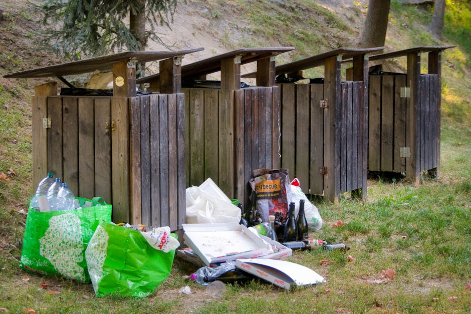Four wooden outdoor waste disposal units with sloped roofs are positioned side by side on a grassy area, each constructed from vertical dark brown wooden planks with visible weathering. In front of the units, a variety of rubbish is scattered across the ground, including a large green plastic shopping bag filled with empty plastic bottles, a white takeout pizza box with some leftover food, and a black bag labeled 'barbecue' containing discarded items. Several empty glass bottles and a few plastic bottles are also visible among the rubbish, alongside some loose packaging and waste. The background features a hillside with a few trees and shrubs, creating a natural setting that contrasts with the visible litter and the functional purpose of the waste units, which appear suitable for private or independent waste disposal outside a typical residential or natural environment. The scene is well-lit, capturing the details of both the waste and the wooden structures clearly, reflecting a situation where rubbish has been improperly disposed of outside designated waste collection points, supporting the theme of alternative rubbish removal services.