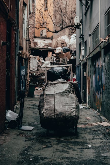 A narrow urban alleyway is cluttered with a large, wheeled black garbage bag in the foreground, partially covered with a grey protective cover. Behind it, there are stacks of discarded cardboard boxes, plastic bags, and mixed waste materials piled against a weathered brick wall and a wooden structure. The alley is flanked by tall, graffiti-covered metal walls on the right and buildings with visible wiring and pipes on the left. In the background, leafless tree branches extend over the waste heap, with a glimpse of a pale, overcast sky. The ground is uneven and stained, reflecting the typical environment of private rubbish clearance sites used by waste removal services like Rubbish Removal Clapham for efficient, independent waste handling in city settings. Overall, the scene highlights a typical cluttered back alley used for waste disposal, emphasizing the importance of reliable rubbish removal.